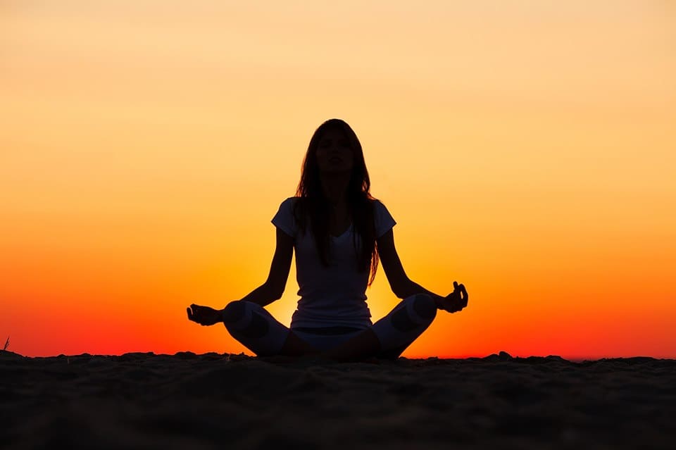 A woman is meditating on the beach at sunset.