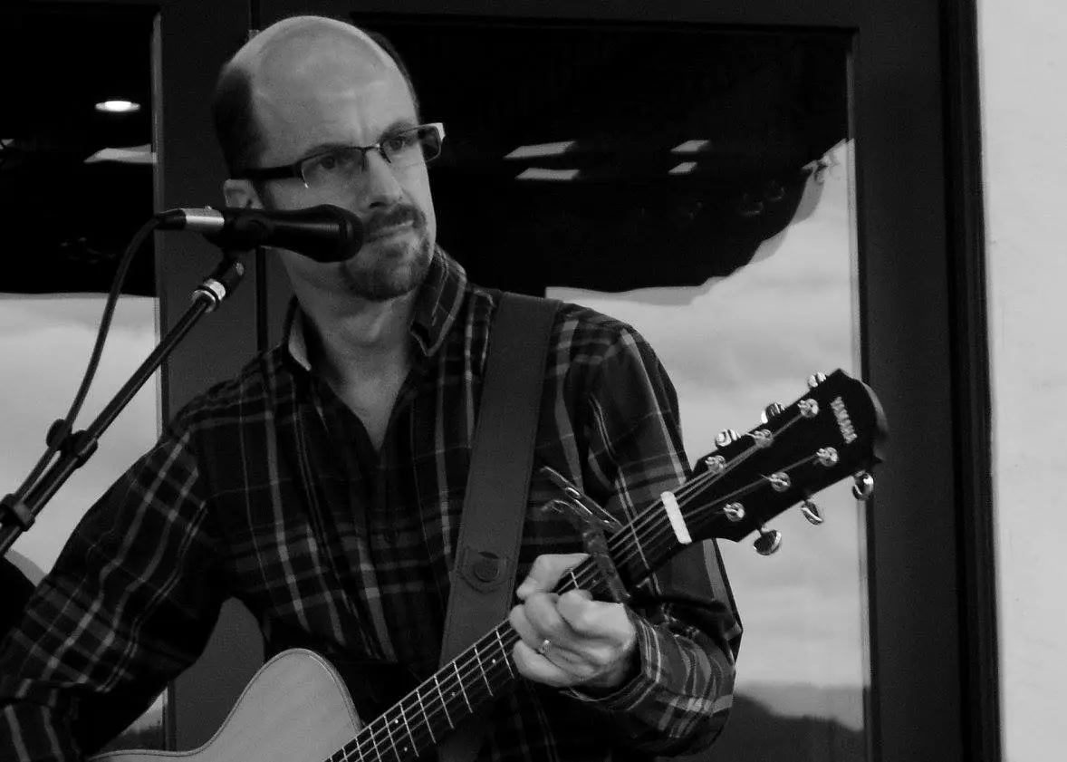 A grayscale image of a man with glasses holding his guitar