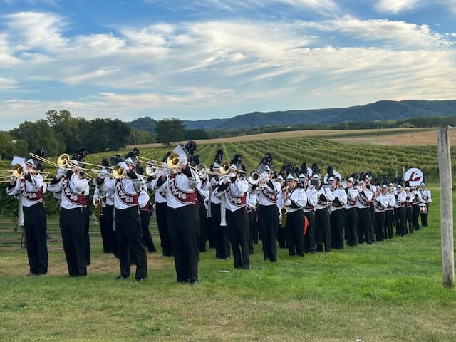 High school marching band plays outdoors in formation with scenic hills.