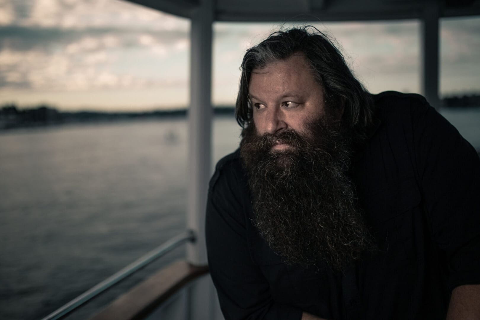 A bearded man gazing thoughtfully over water on a boat.