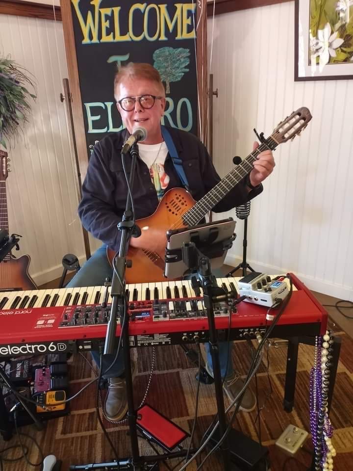 Man playing guitar and keyboard, smiling at the camera during a live music session.