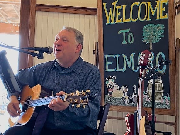 Man playing acoustic guitar and singing at a venue with a welcome sign.