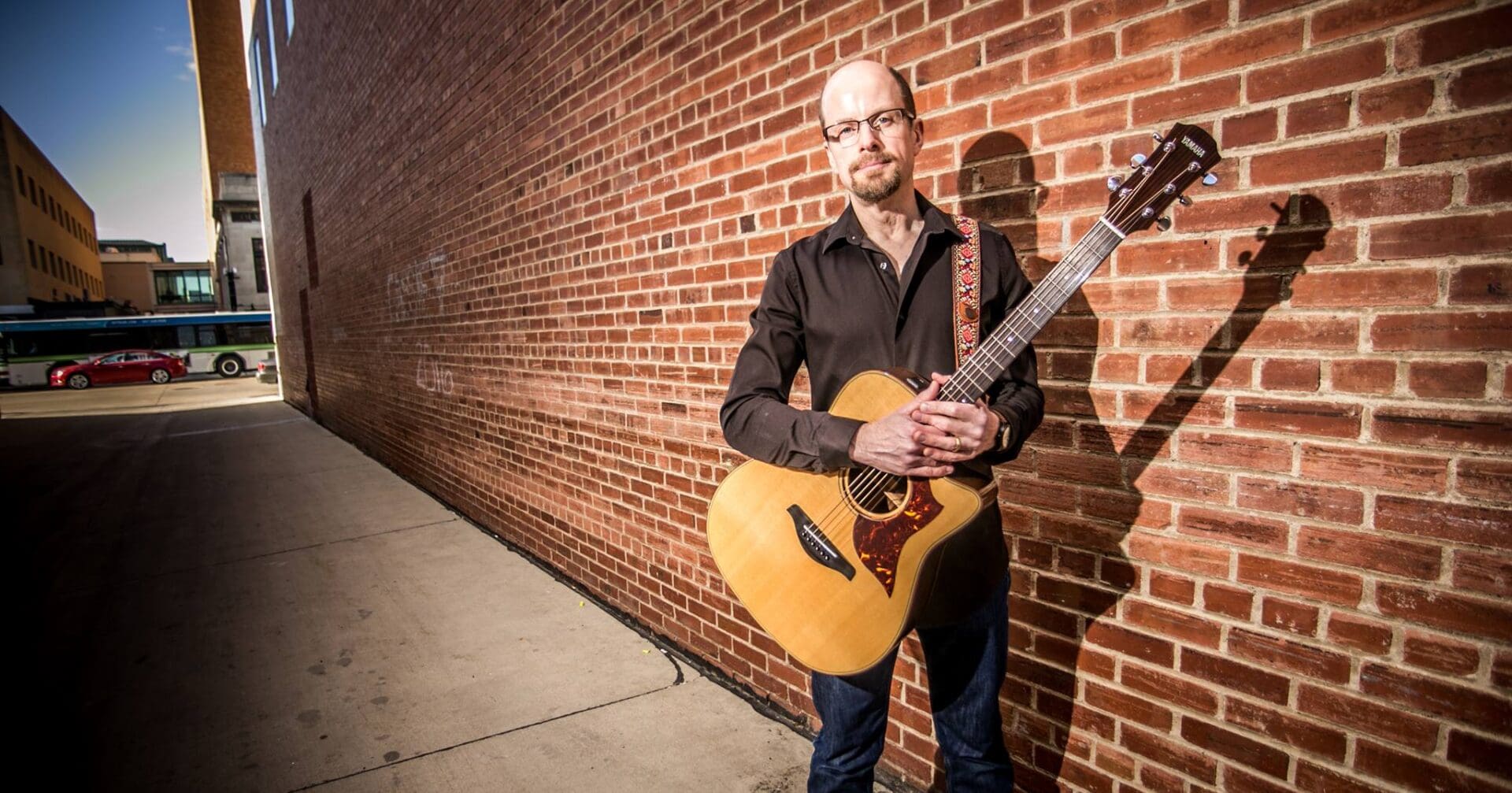 Man standing against brick wall holding an acoustic guitar.