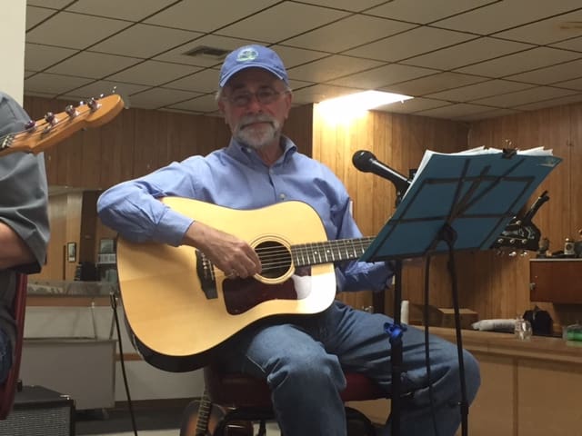 Older man playing acoustic guitar at a casual indoor gathering.