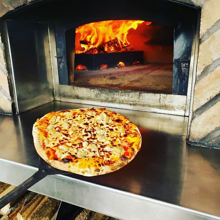 A freshly baked pizza coming out of a wood-fired oven with flames inside.