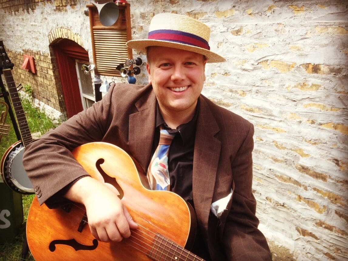 Smiling man in vintage attire playing an acoustic guitar outdoors.