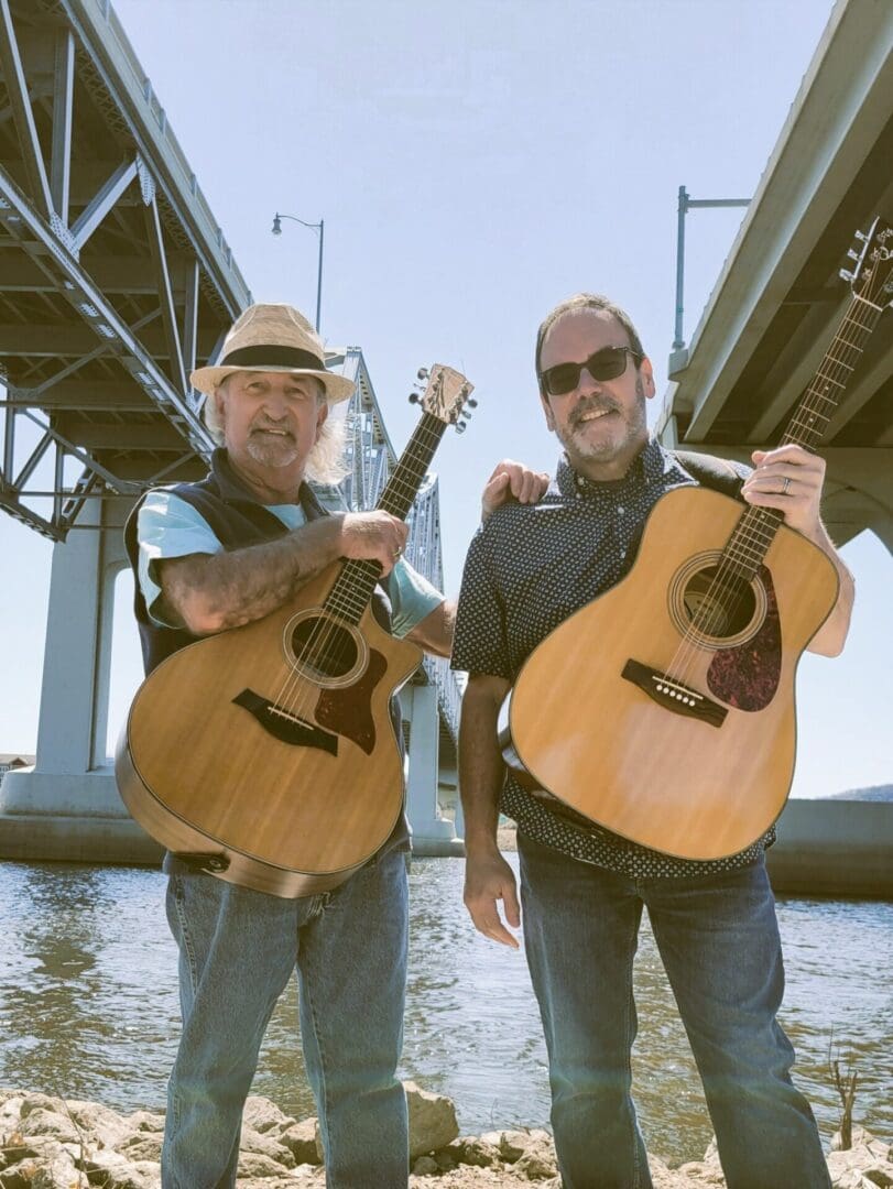 Two men with guitars standing by a river under a bridge on a sunny day.