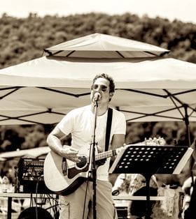 A man passionately singing and playing guitar outdoors under a canopy.